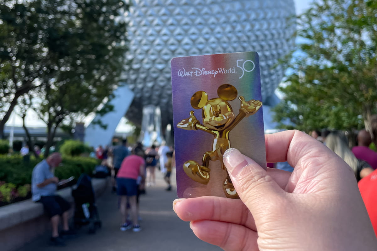 Close up of a person's hand holding a Disney World Annual Pass with the EPCOT sphere in the background