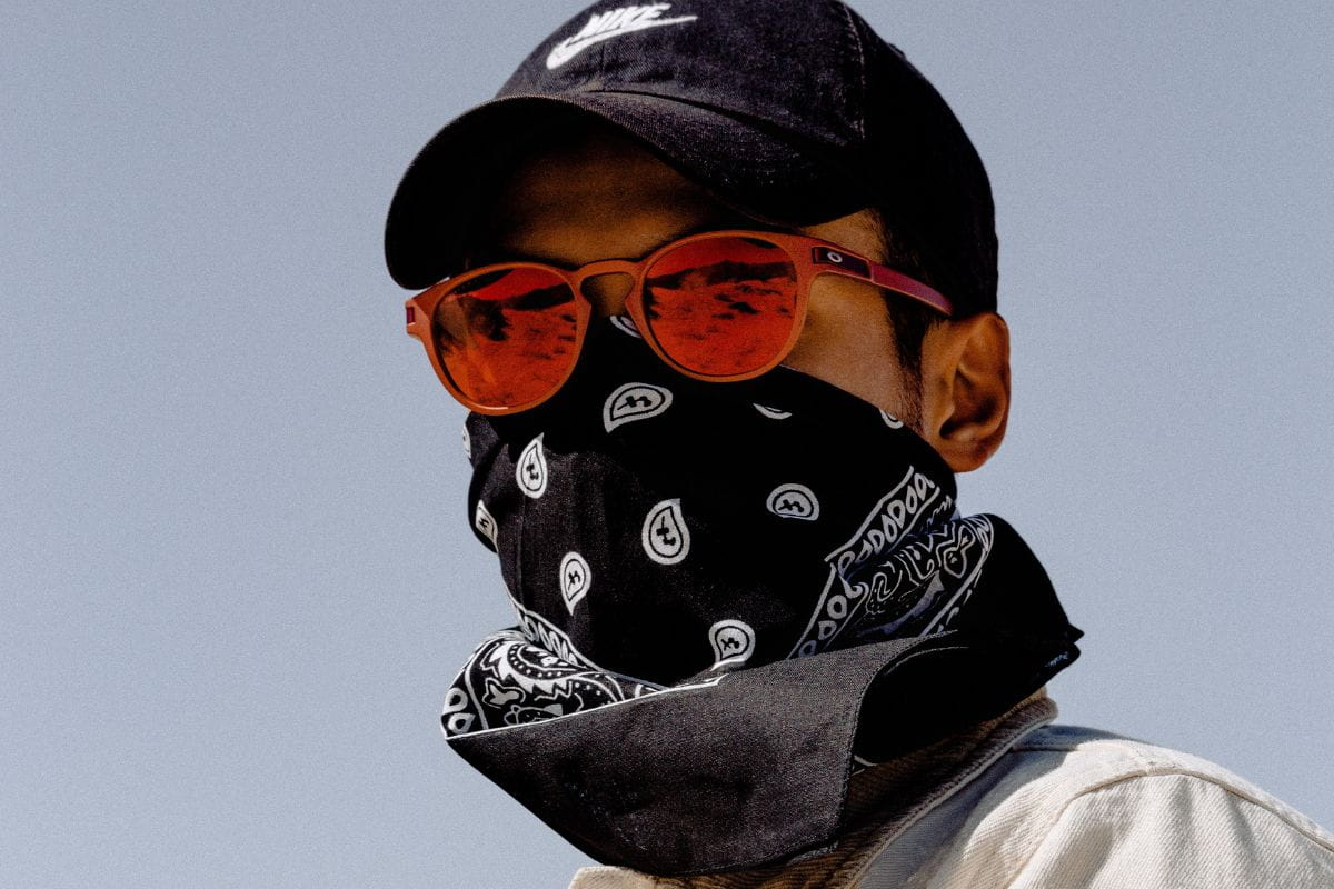 Person wearing a black bandana covering the lower half of face, orange shades, and a black cap