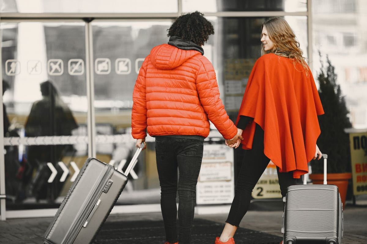Woman wearing an orange poncho and a man wearing a puffer jacket both holding suitcases