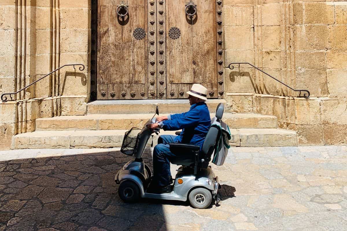 Person riding a mobility scooter passing by a stone building with a wooden door