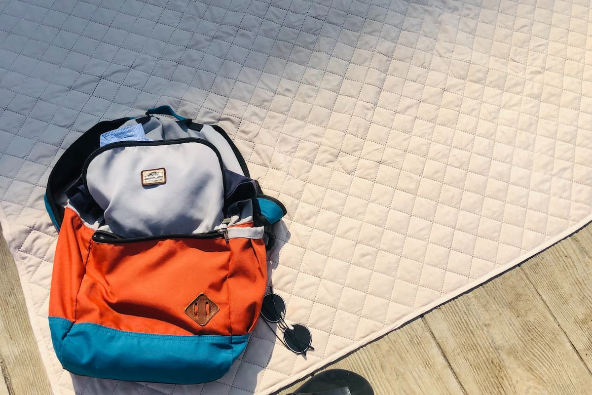 Flat lay photo of a blue, orange, and gray backpack on the floor with sunglasses beside it. 