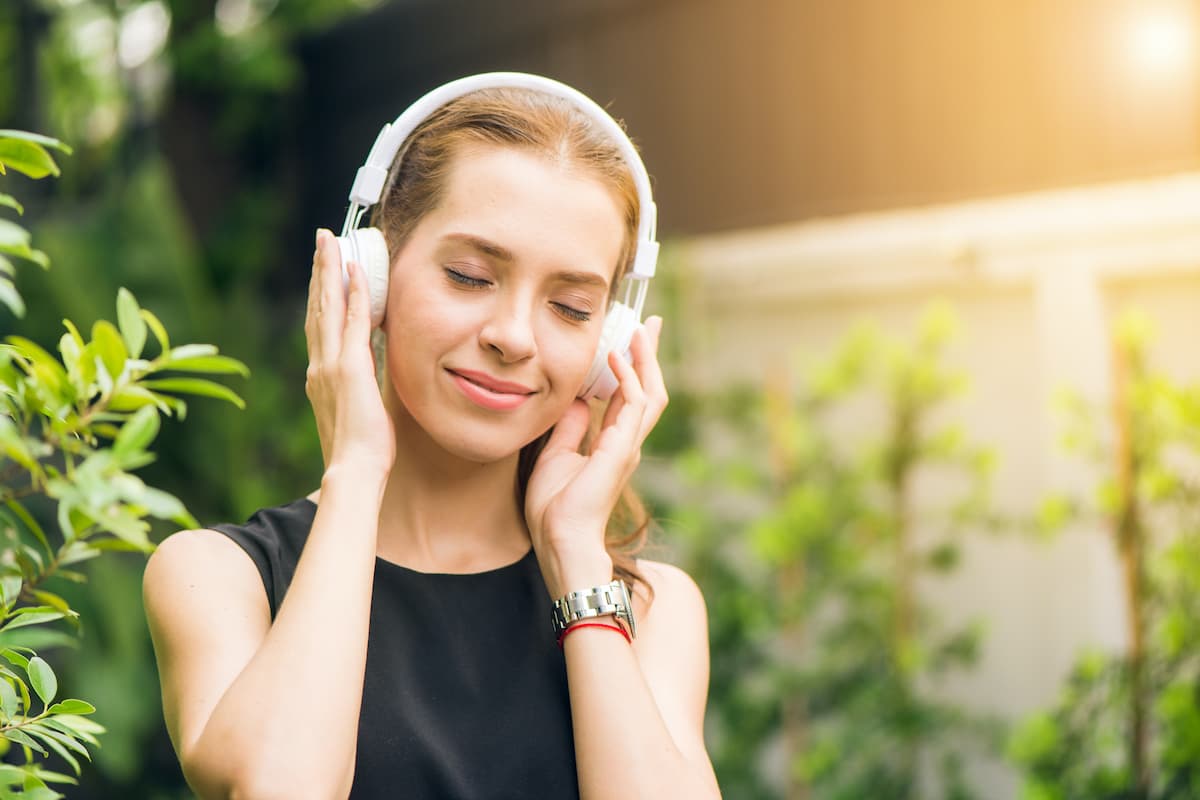 A woman in a black sleeveless shirt is holding white headphones while listening to music.