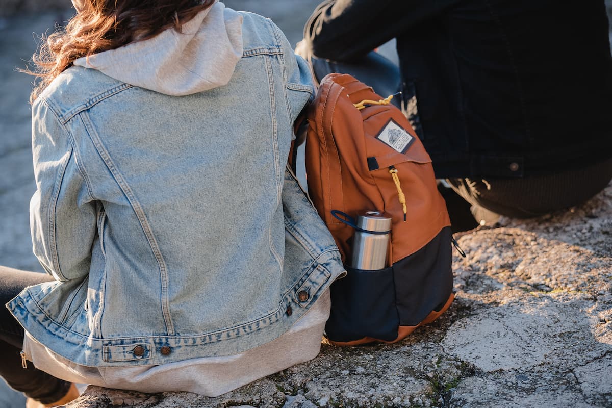 Cropped photo of a woman in a denim jacket and a man in a black jacket sitting on a rock with a brown backpack with a silver tumbler between them.