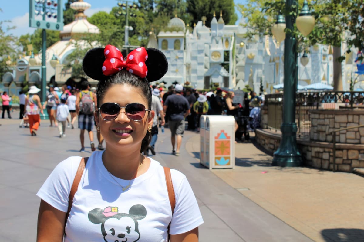 A woman is wearing sunglasses, Minnie ears, and a white t-shirt with Minnie Mouse printed on it. 