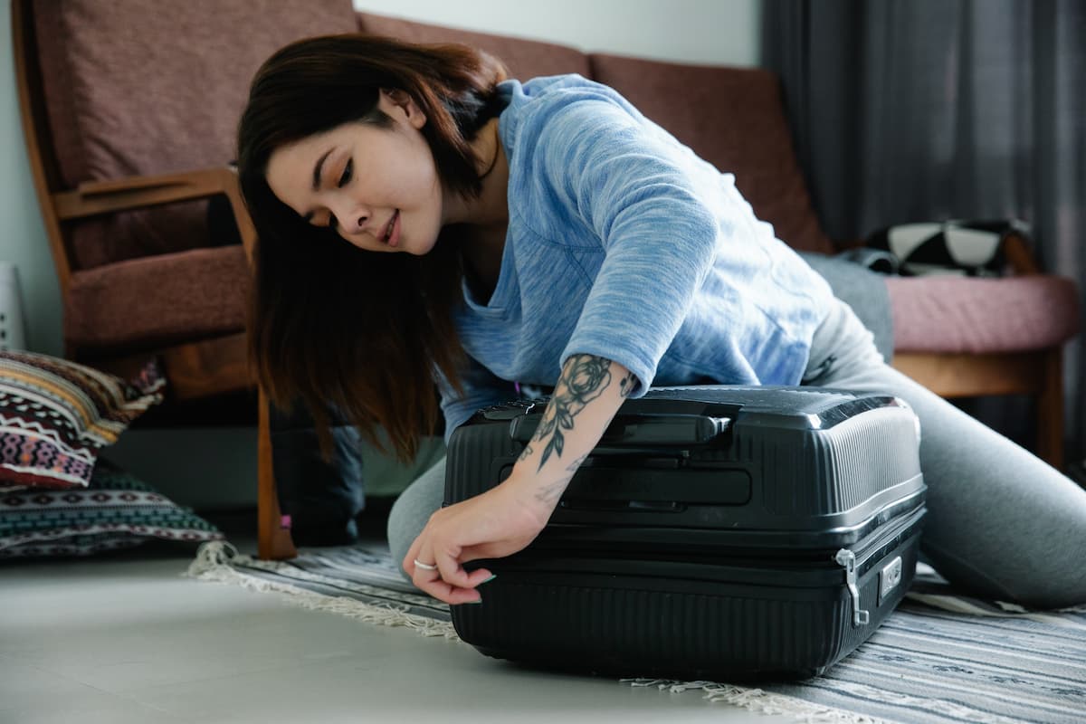 A woman in a blue sweatshirt is zipping a suitcase on the floor. 