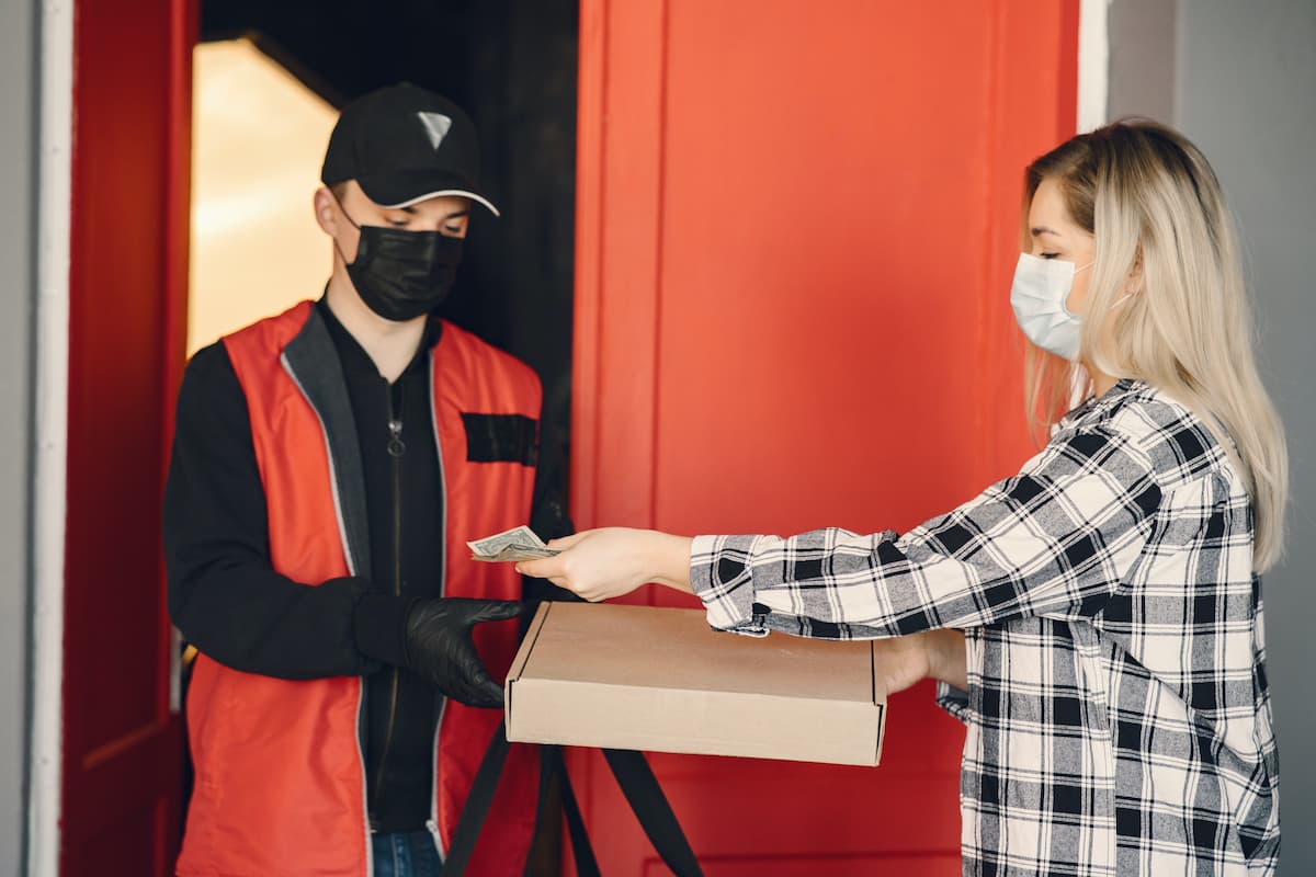 A woman wearing a mask pays the delivery man for the pizza. 